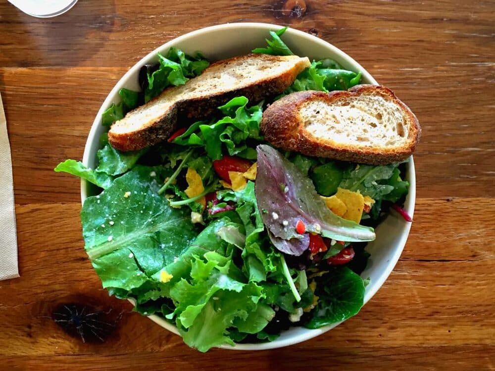 A bowl of leaves, tomato and toasted bread on a wooden table.