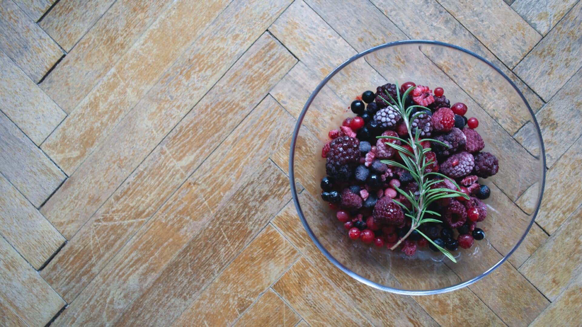 A glass bowl of blackberries, raspberries and redcurrants with a sprig of rosemary, on a pale herringbone wood floor.
